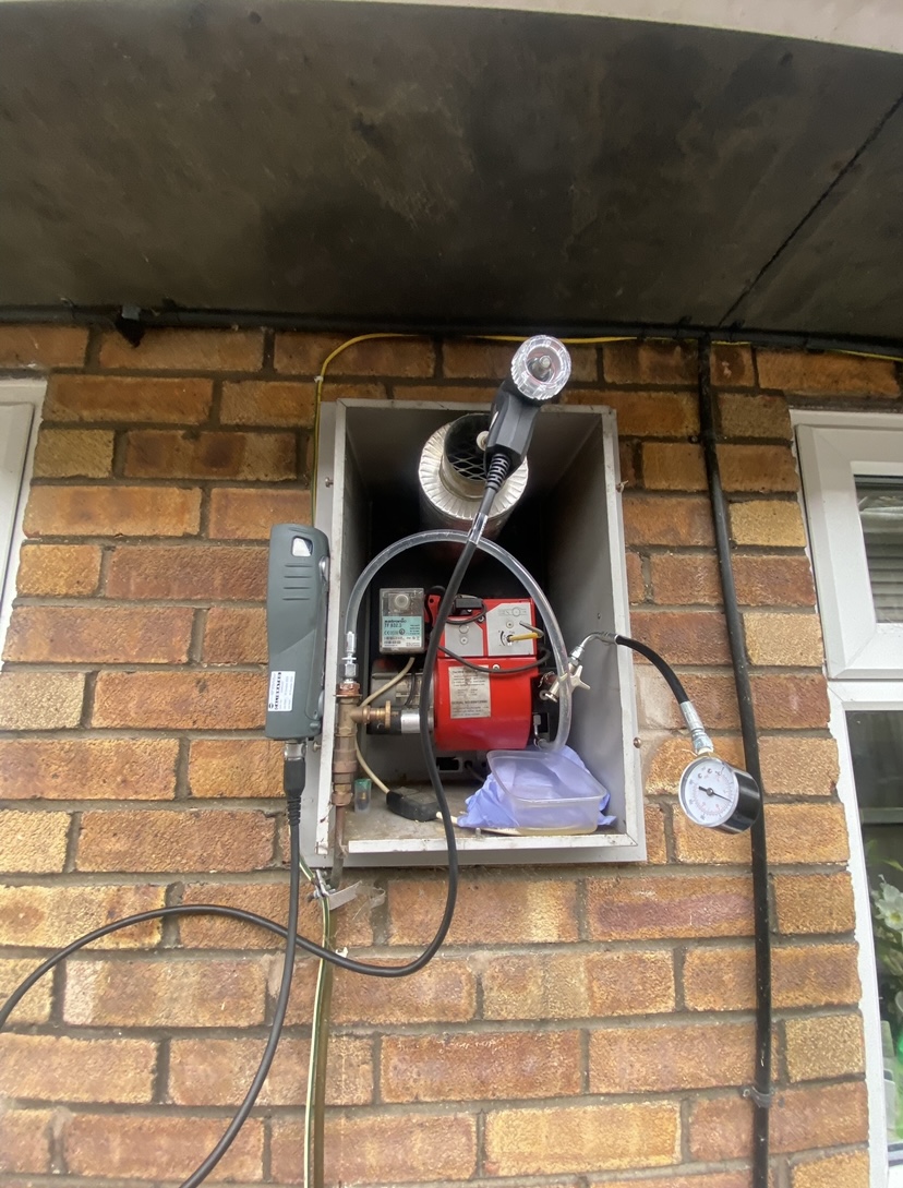 An outdoor oil burner on a brick wall with a pressure gauge connected for testing